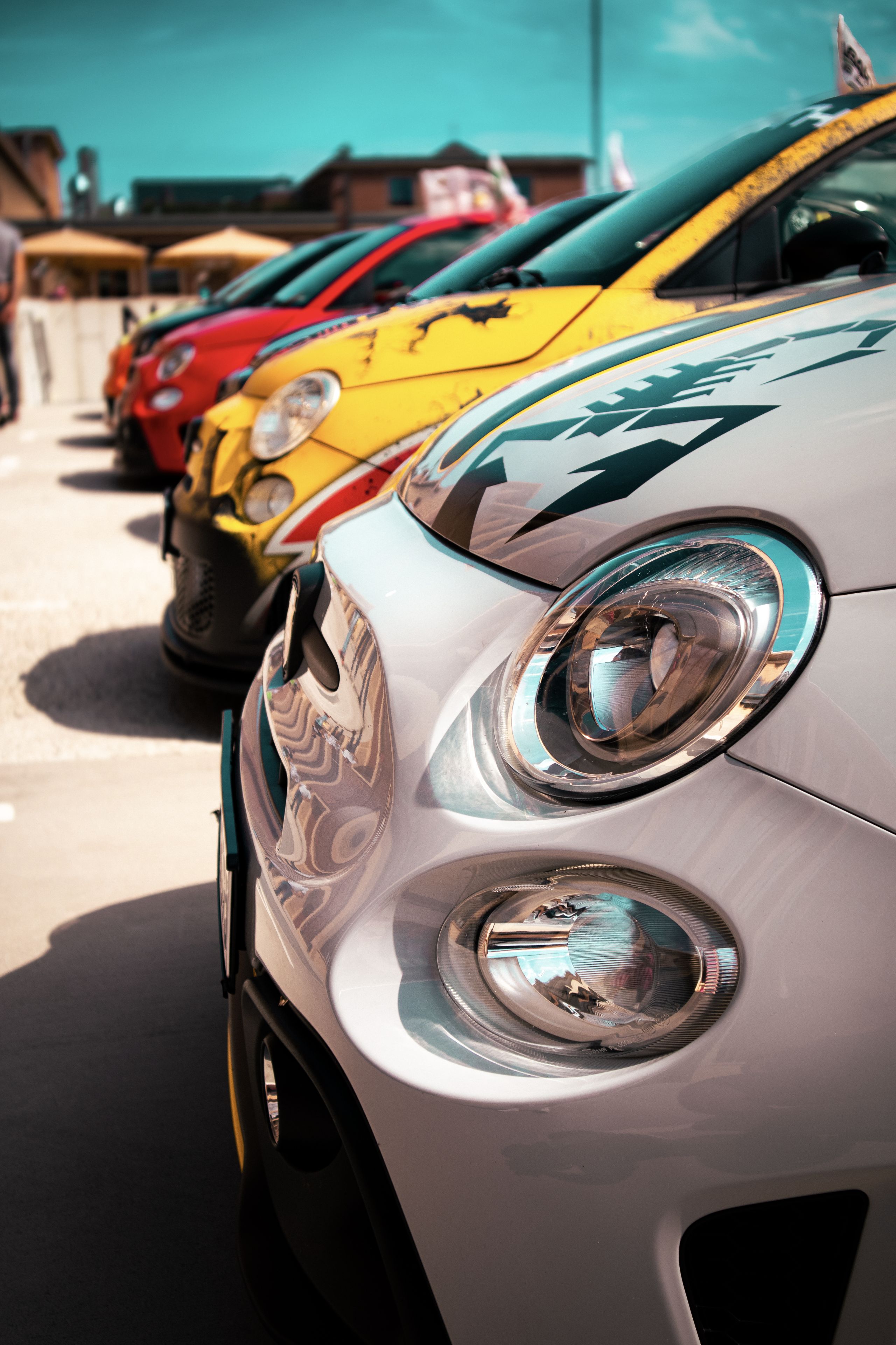 A row of colorful cars, including white, yellow, and red, parked closely together in a sunny outdoor setting