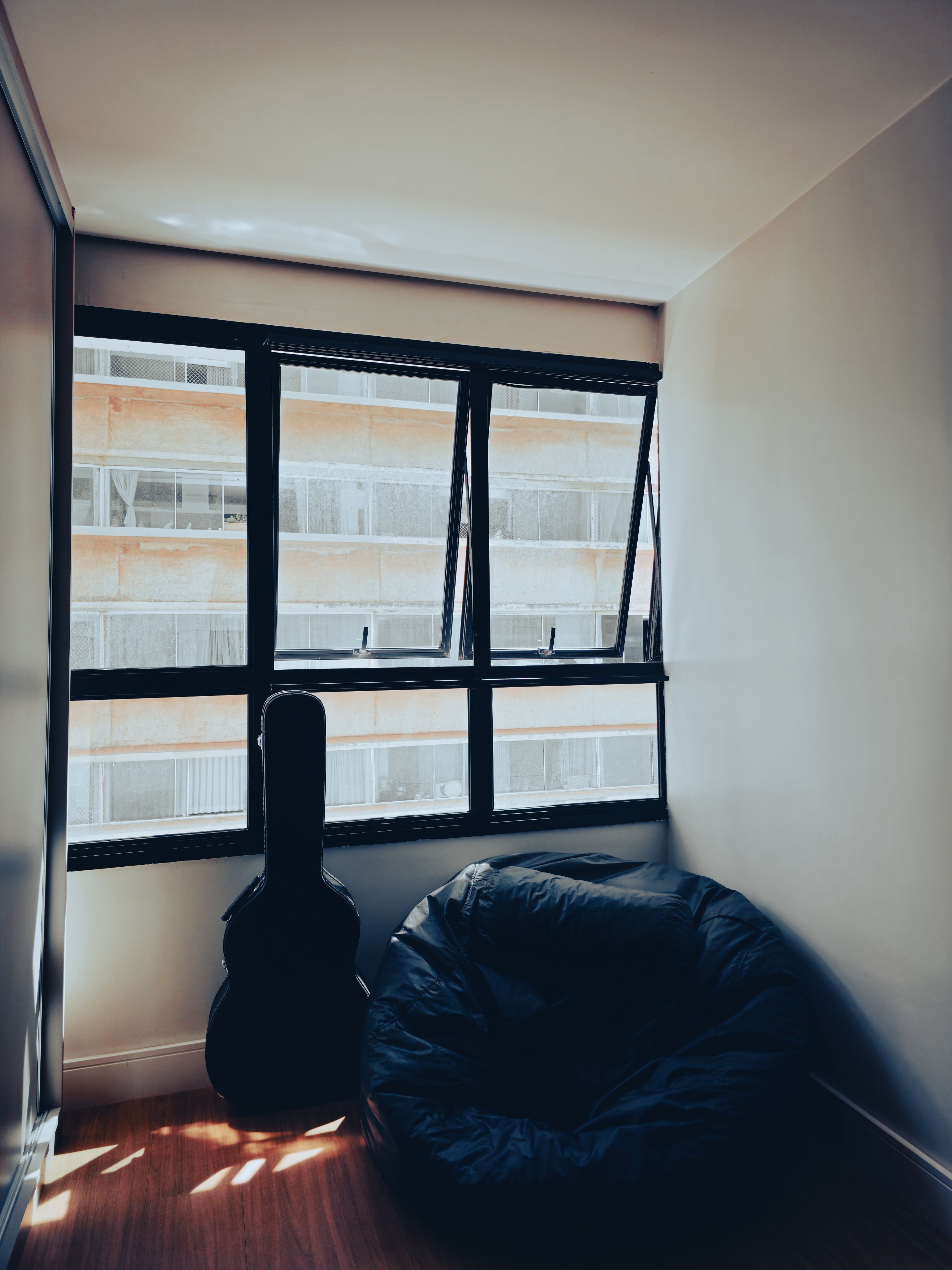 A small room with a large window, a guitar case leaning against the wall, and a bean bag chair on the floor