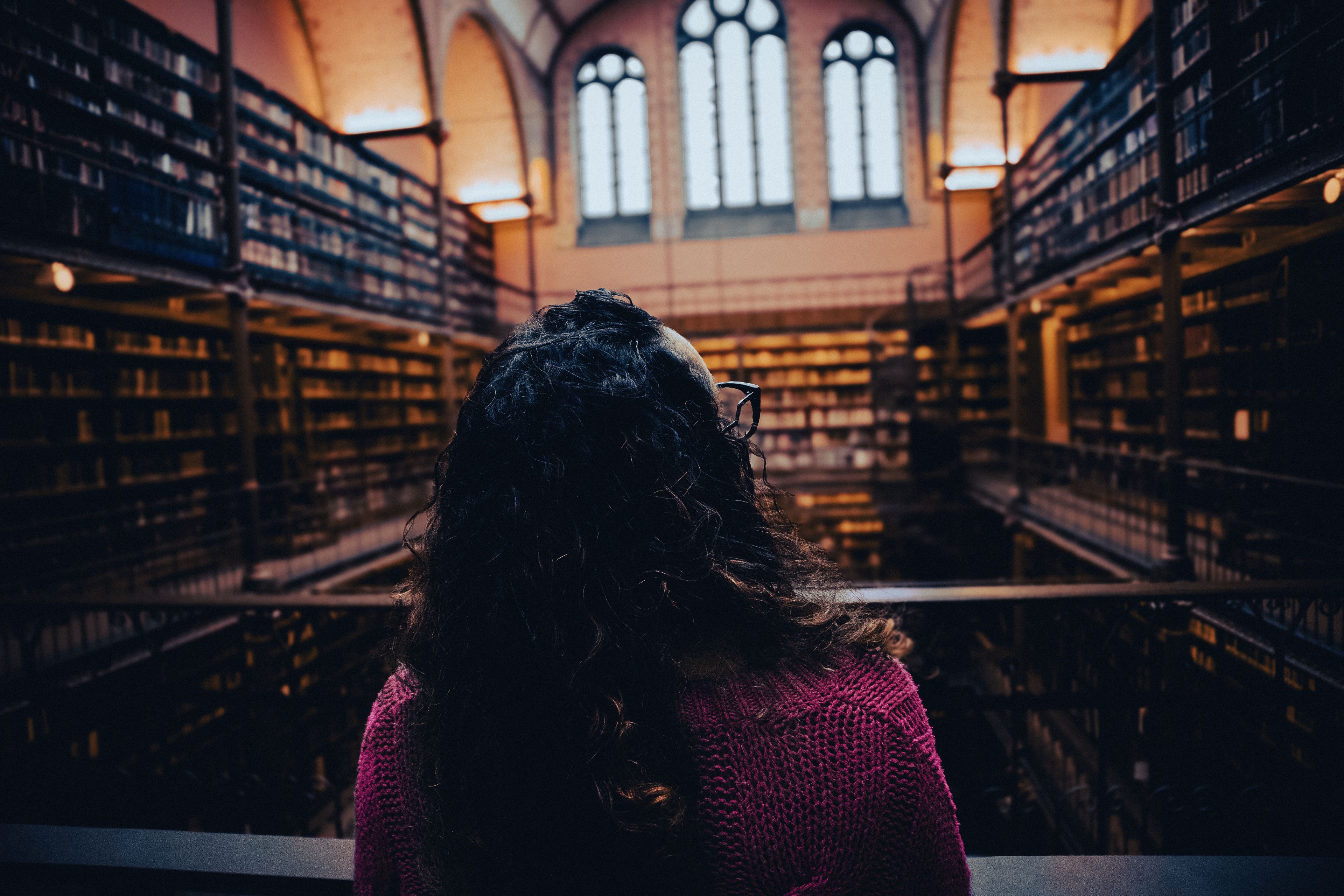 A person with long hair stands in a large, dimly lit library, facing rows of bookshelves and arched windows above