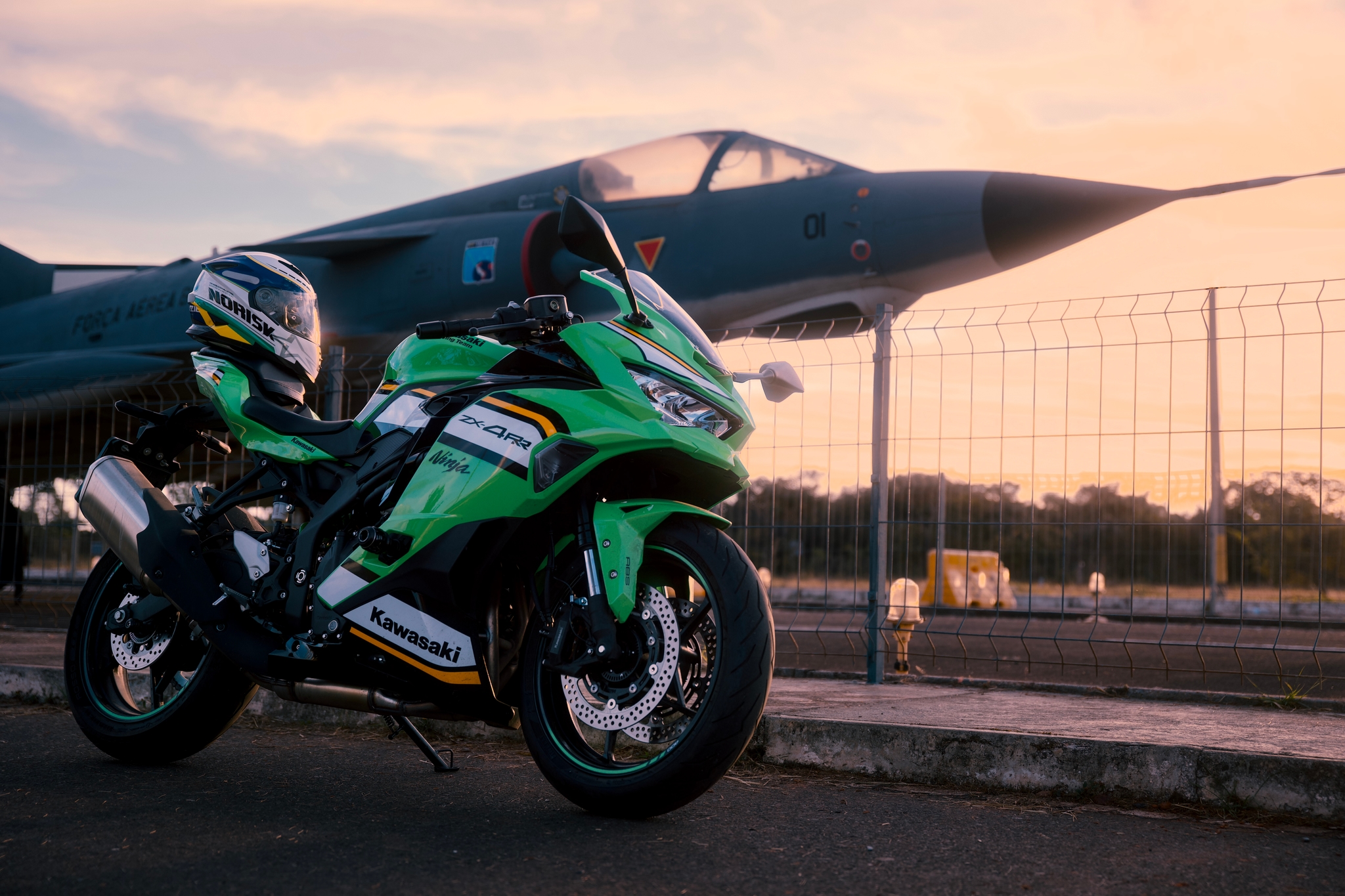 Green motorcycle parked in front of a fighter jet with a sunset backdrop
