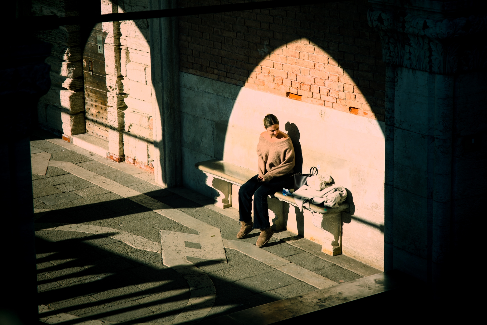 Person sitting on a stone bench beneath an arched passageway, lit by sunlight with long shadows across the walkway