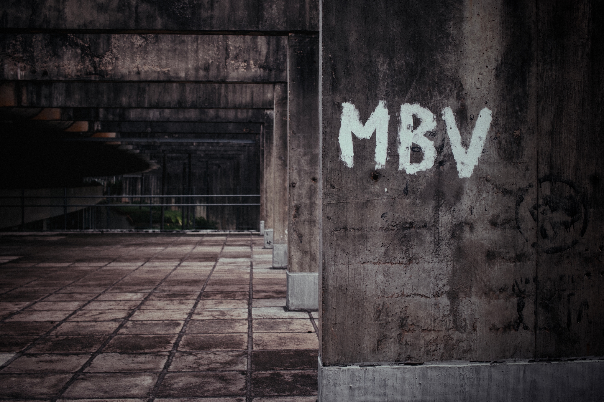 A weathered concrete structure with MBV graffiti on a wall, featuring a tiled floor and a dimly lit, industrial atmosphere