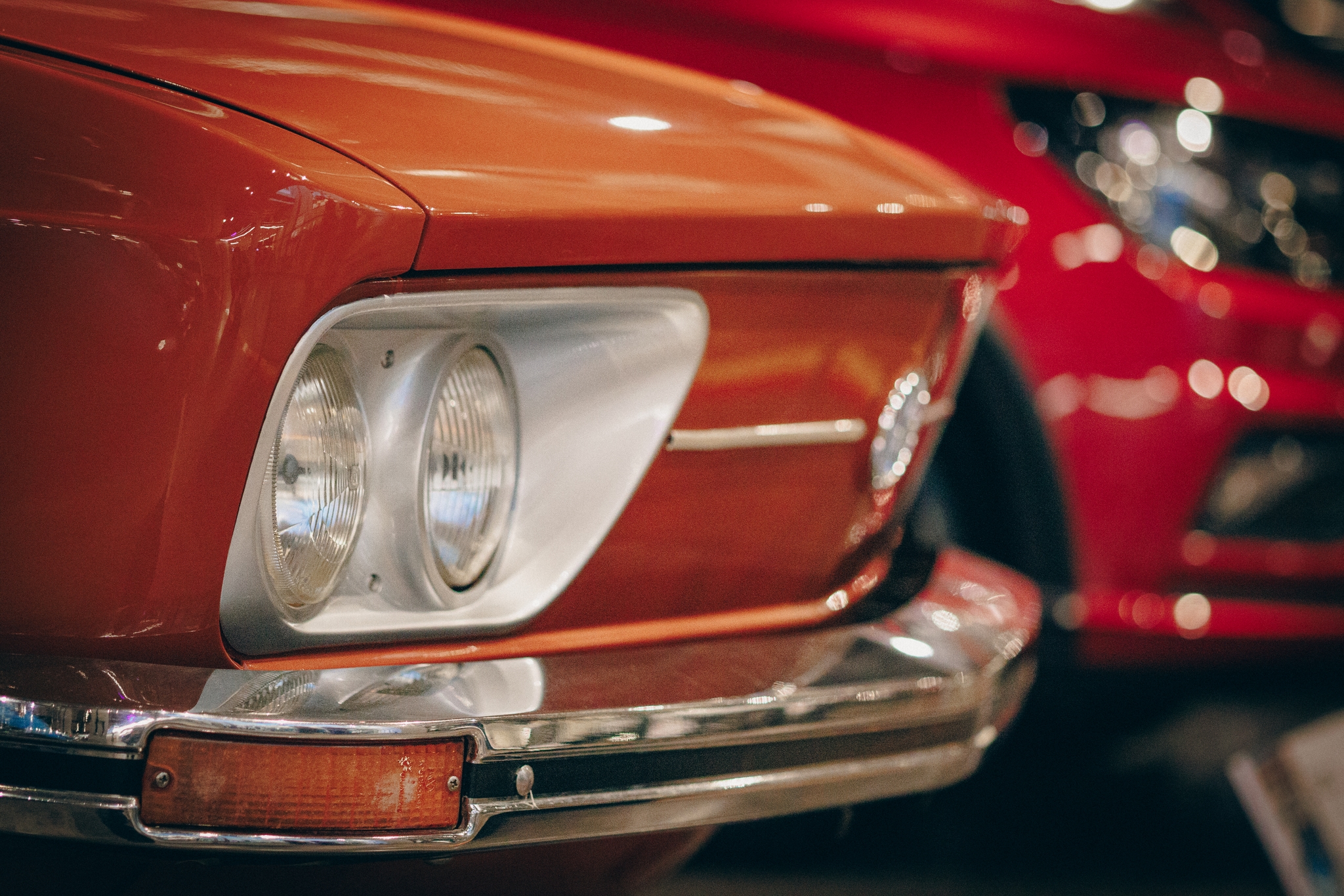 A close-up of the front end of an orange vintage car, focusing on the headlight and grille, with a red car blurred in the background