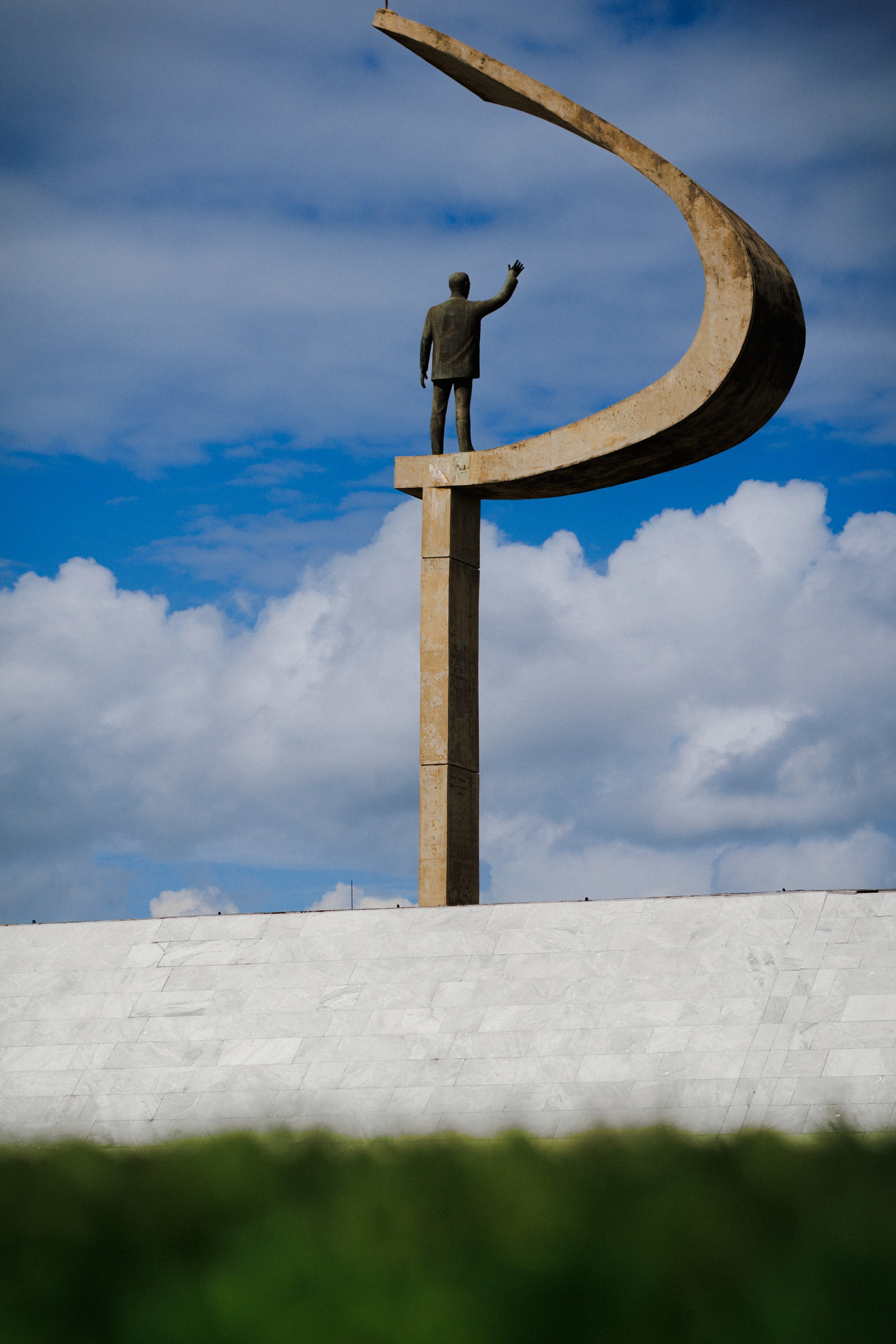 A tall statue of a person standing on a pedestal with an abstract, curved structure above, set against a cloudy sky