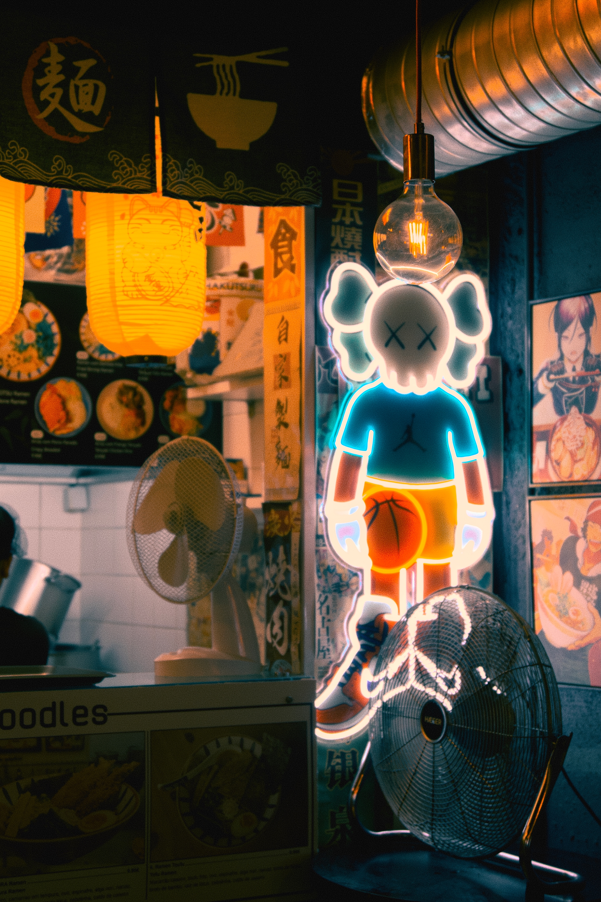 A neon sign of a cartoon character with crossed-out eyes is illuminated in a dimly lit restaurant, surrounded by various food-themed decorations and a fan