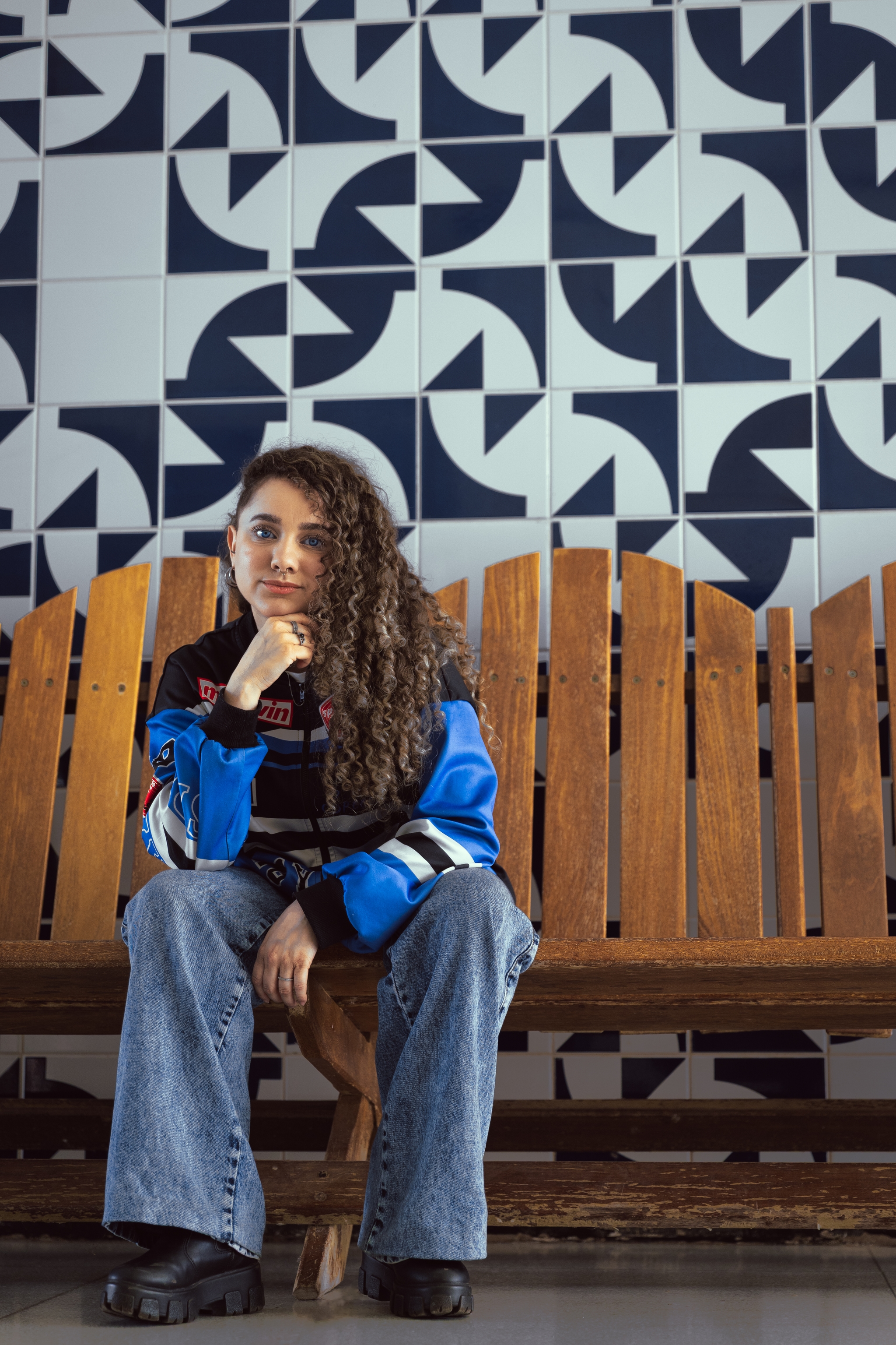 A person with curly hair sits on a wooden bench, wearing a blue and black jacket and jeans, in front of a wall with a geometric black and white pattern