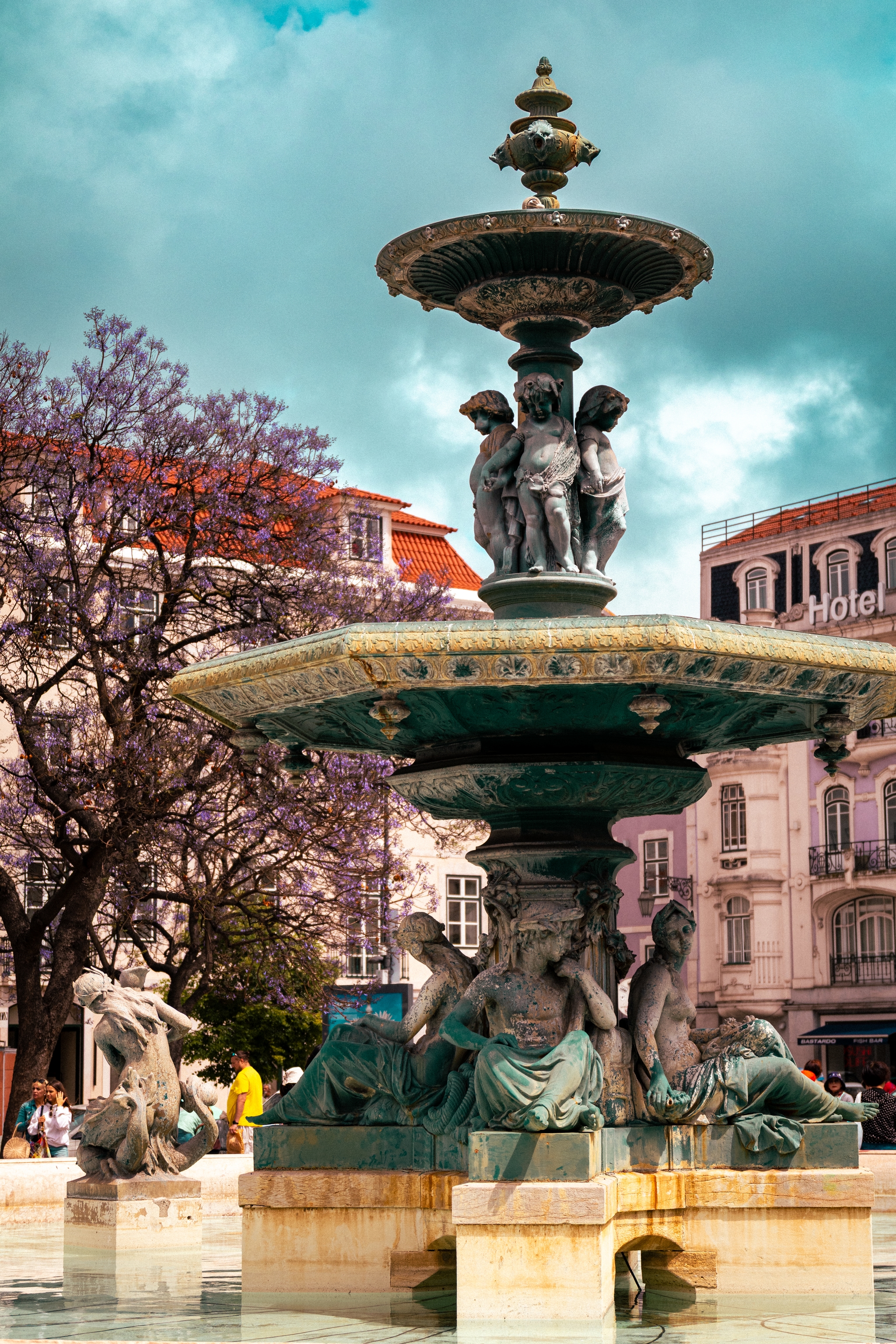 A decorative fountain with intricate sculptures, surrounded by trees and buildings under a cloudy sky