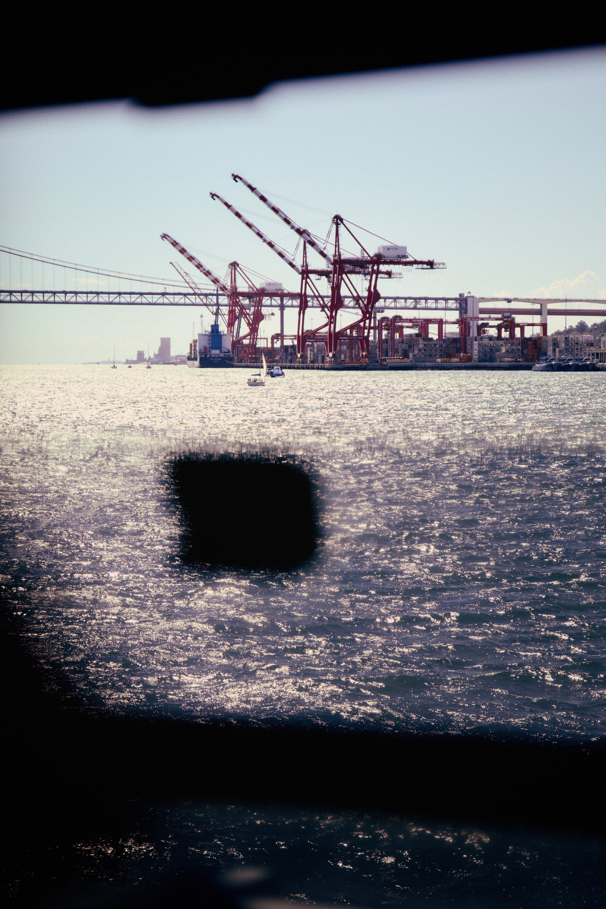 A waterfront scene with large red cranes, a bridge in the background, and sunlight reflecting on the water