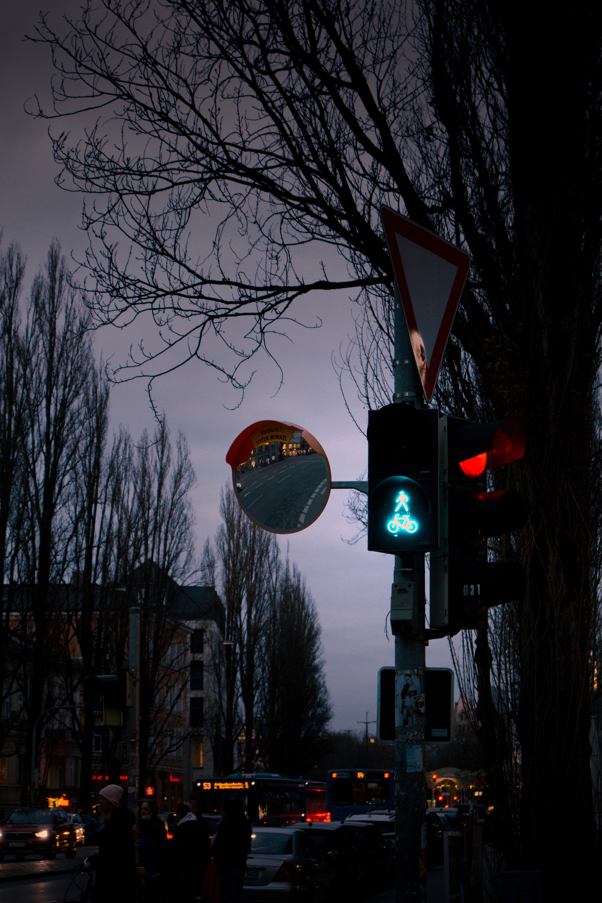 A dimly lit street scene at dusk with a traffic light showing a green pedestrian signal, surrounded by bare trees and a convex mirror reflecting the street