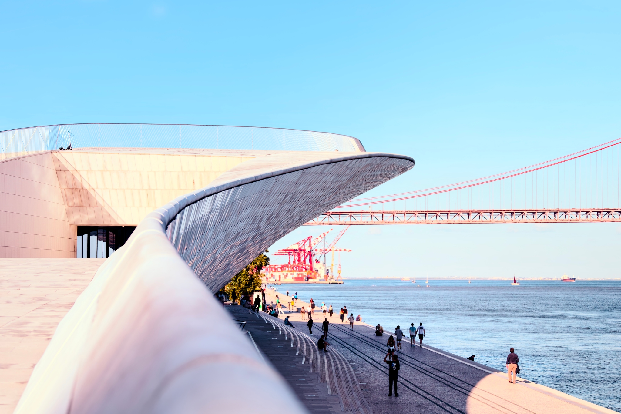 A modern architectural structure with a sweeping, curved design is situated by a waterfront People are walking along the promenade, and a large bridge spans the background under a clear blue sky