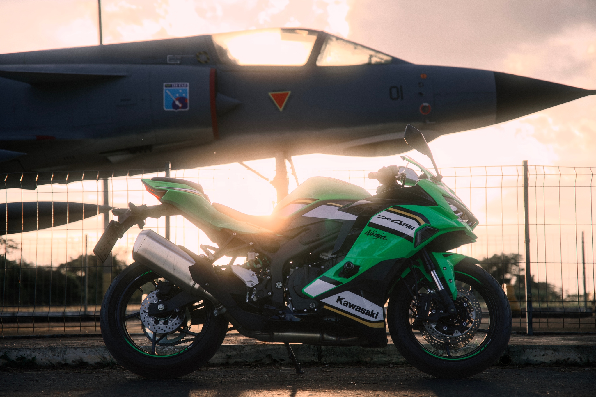 A green motorcycle parked beneath a fighter jet with the sun setting in the background