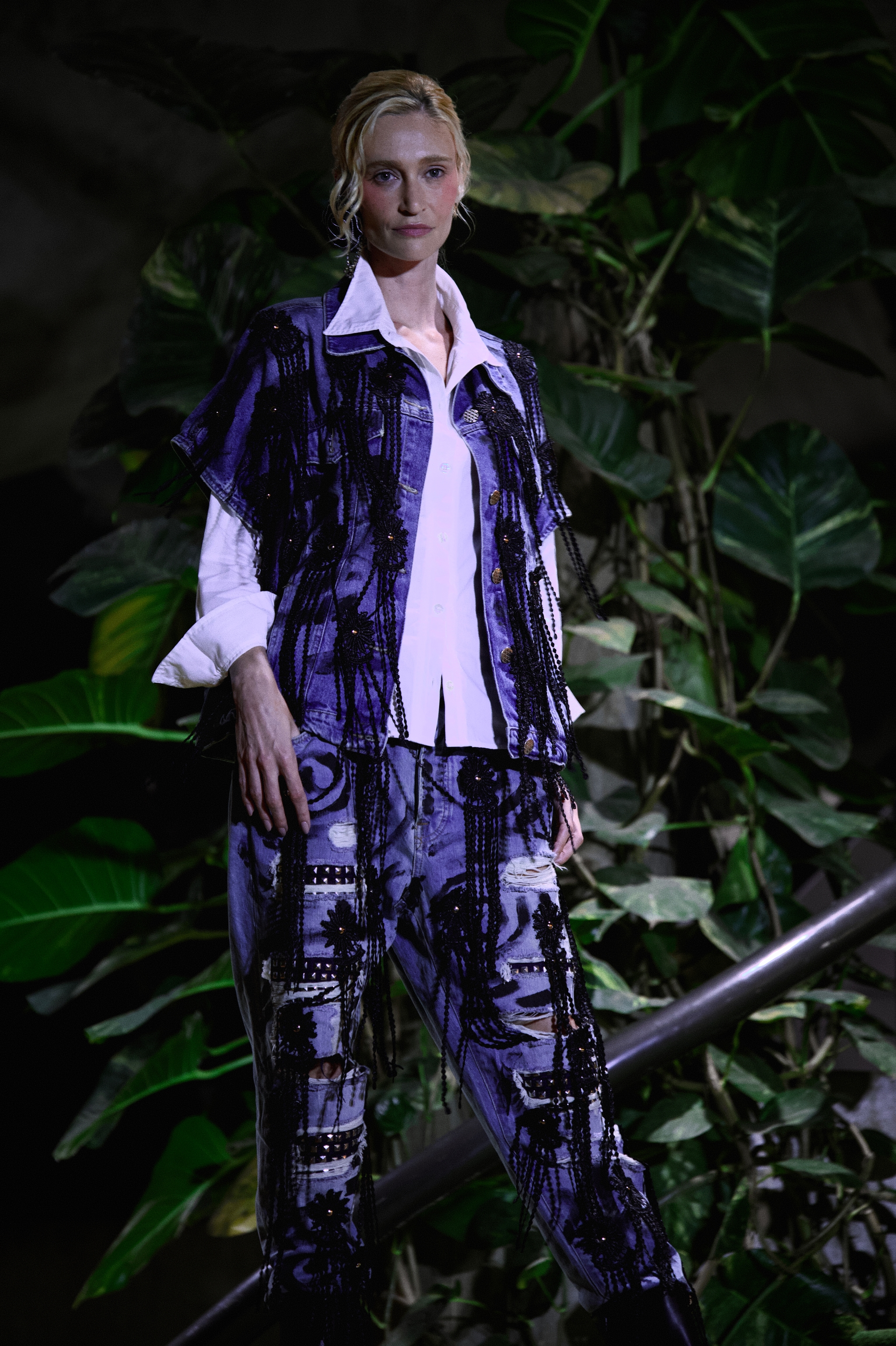 Woman posing in a patterned outfit over a white shirt on a stairway with leafy plants in the background under low light