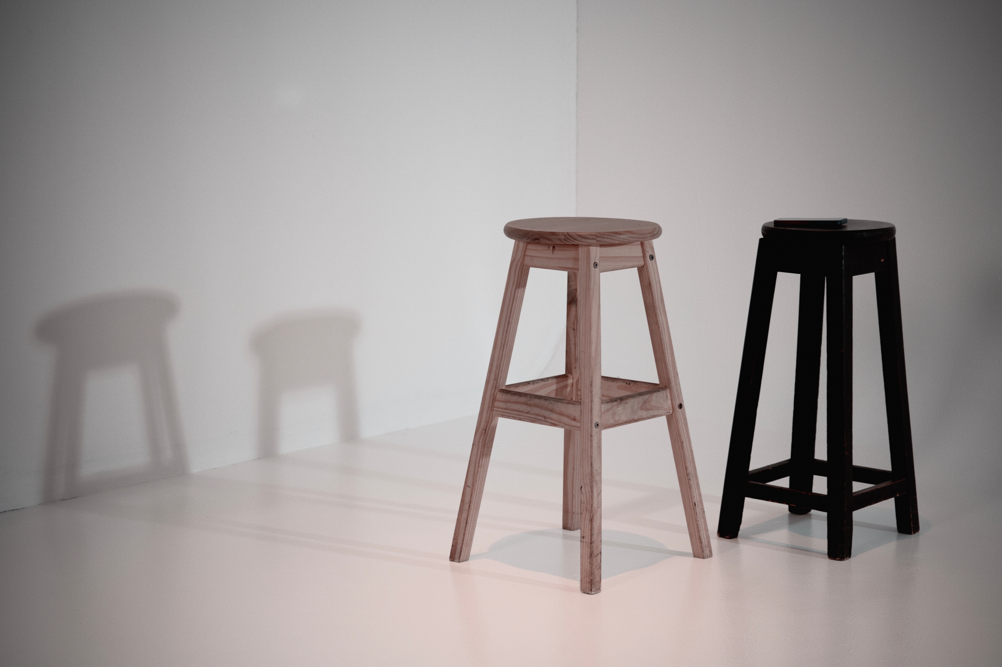 Two stools, one light brown and one black, are placed on a white surface with their shadows cast on the wall behind them