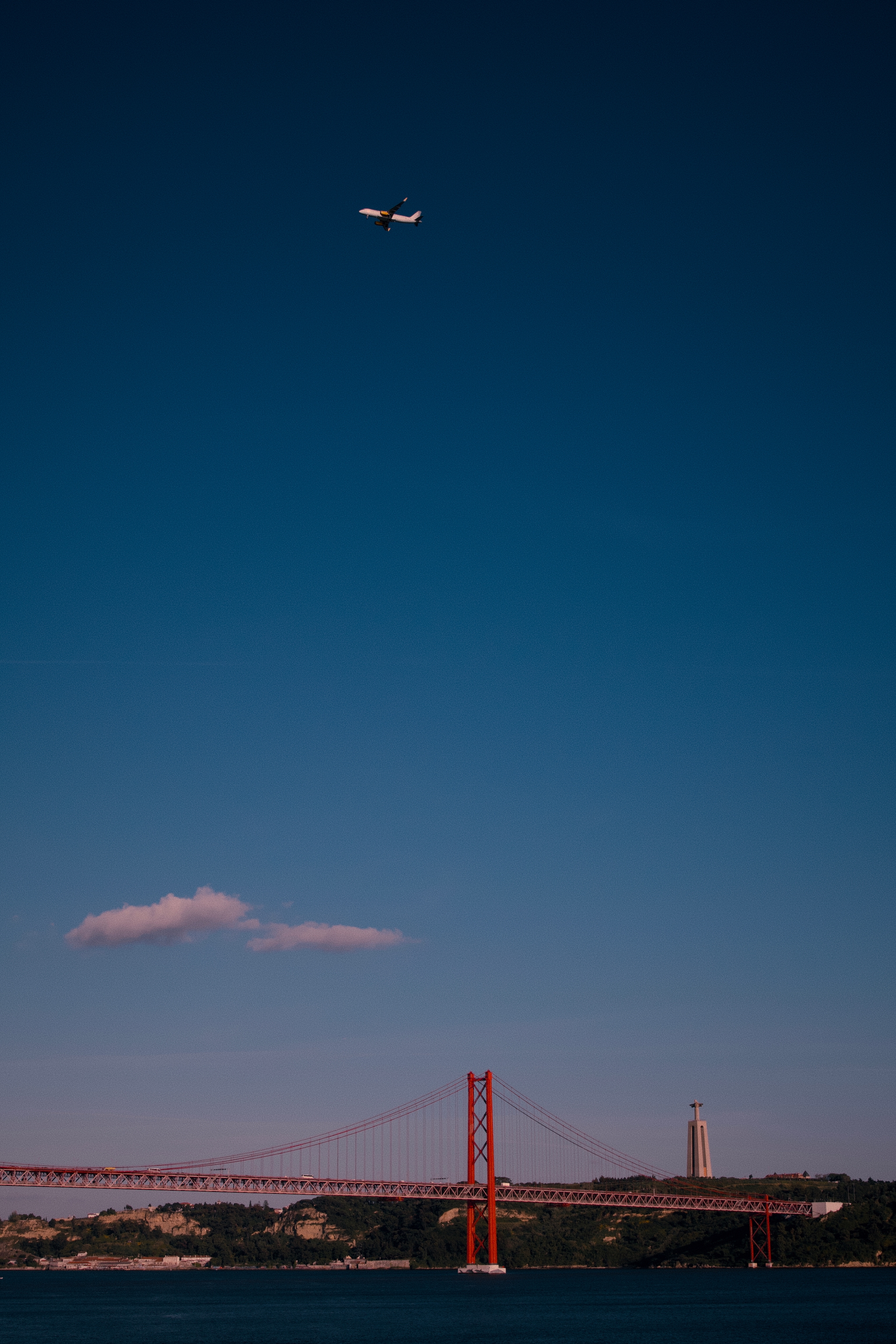 A red suspension bridge spans across a body of water with a plane flying above in a clear blue sky