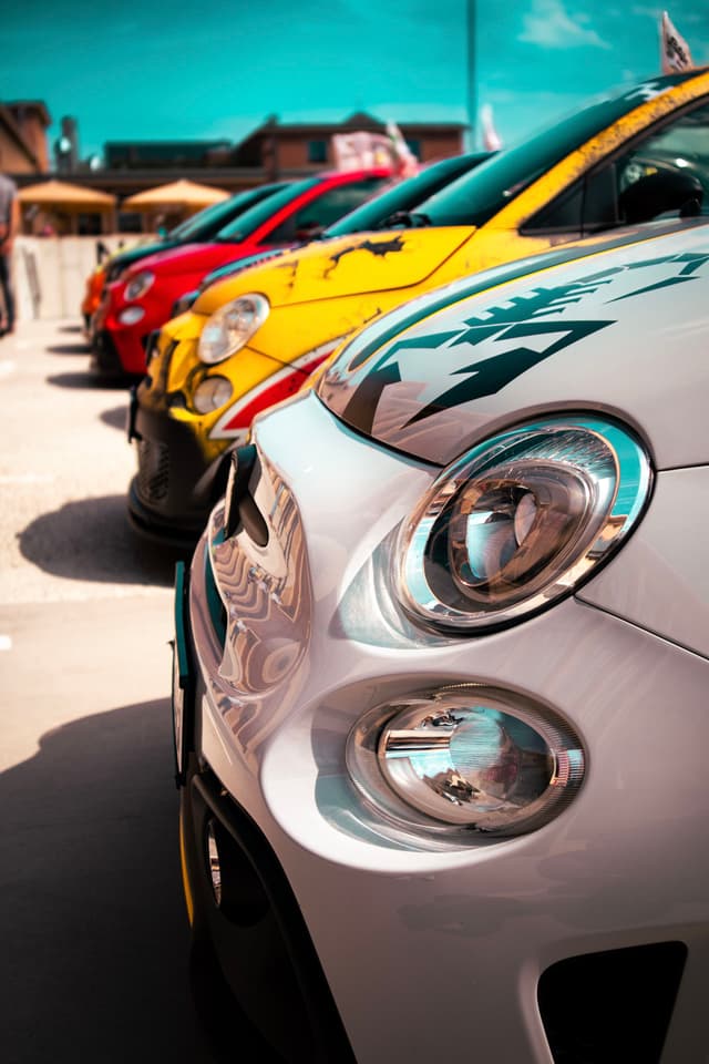 A row of colorful cars, including white, yellow, and red, parked closely together in a sunny outdoor setting