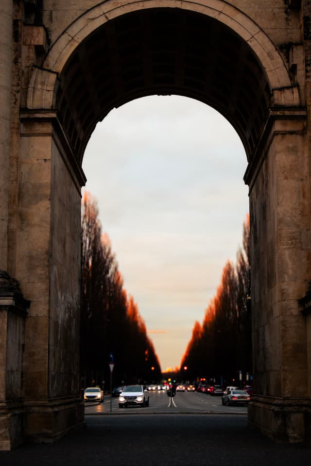 A large stone archway frames a view of a straight road lined with trees, with cars and a sunset sky in the background