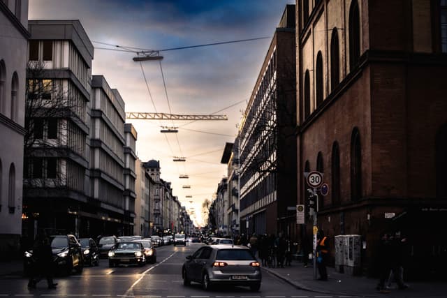 A city street at dusk with cars and pedestrians, flanked by tall buildings. Overhead, streetlights are suspended, and the sky is a mix of blue and orange hues
