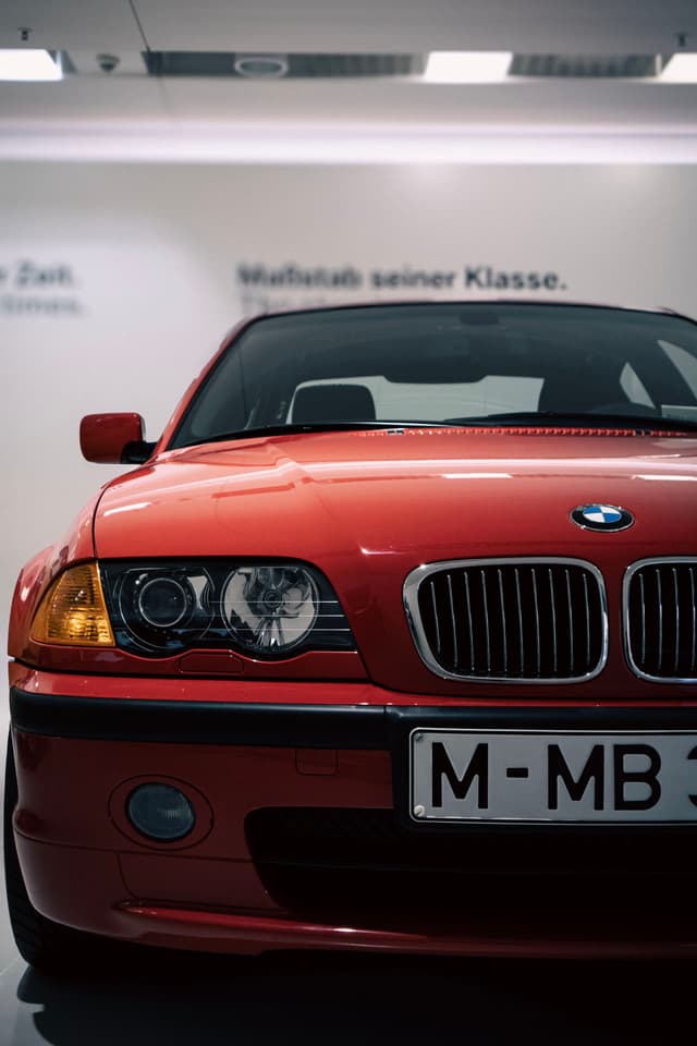 A red BMW car is displayed indoors, with a focus on the front grille and headlights. The background features blurred text on the wall