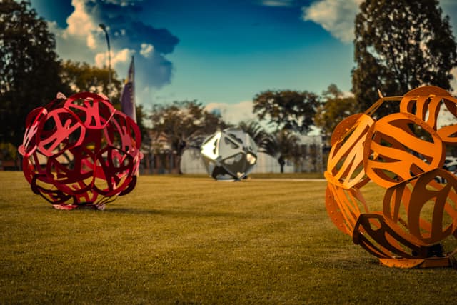 Three large, colorful geometric sculptures are placed on a grassy field, with trees and a partly cloudy sky in the background