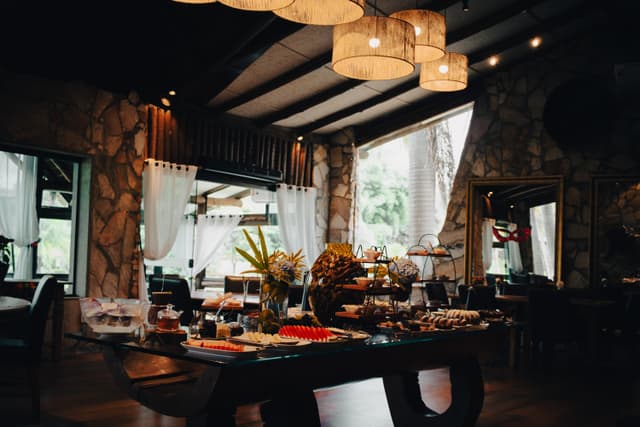 A cozy restaurant interior with stone walls, large windows, and a table set with various dishes and decorations. Warm lighting and elegant furnishings create an inviting atmosphere