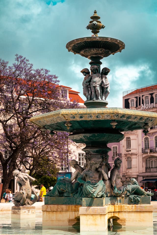 A decorative fountain with intricate sculptures, surrounded by trees and buildings under a cloudy sky