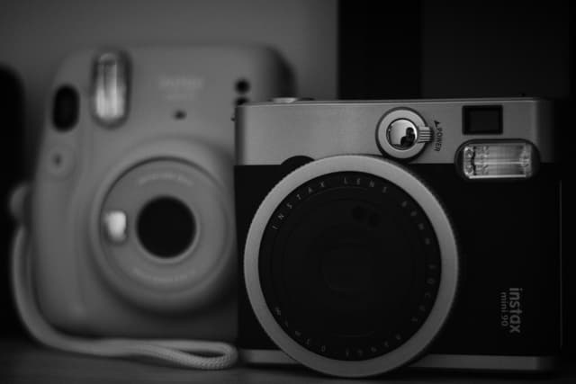 Two vintage-style instant cameras, one in the foreground and one slightly blurred in the background, captured in black and white