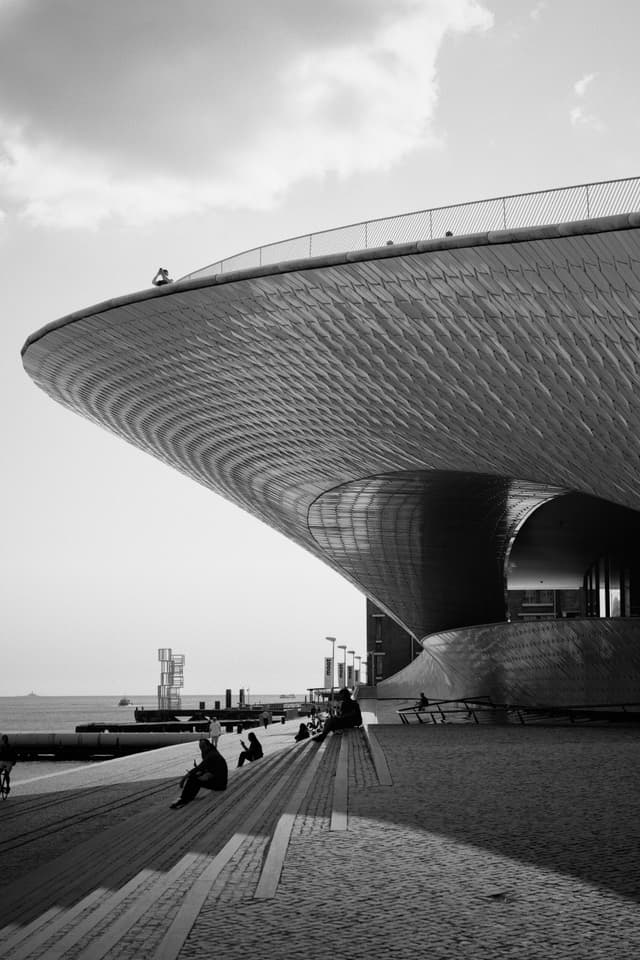 A black and white photograph of a modern architectural structure with a sweeping, curved roof People are sitting on steps in the foreground, and the scene overlooks a waterfront with a cloudy sky