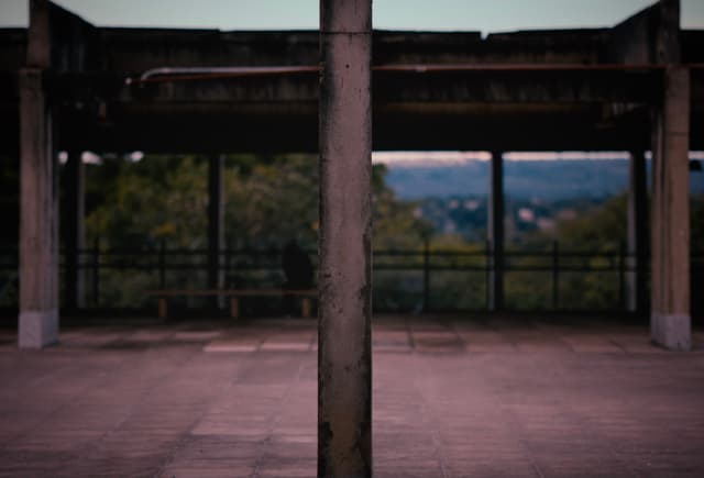 A concrete pillar stands in the center of an open, empty structure with a view of trees and a distant landscape in the background