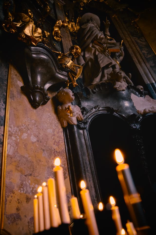 Lit candles in the foreground before an ornate, shadowed altar with carved figures and decorative stonework inside a dim interior