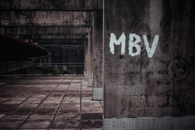 A weathered concrete structure with MBV graffiti on a wall, featuring a tiled floor and a dimly lit, industrial atmosphere