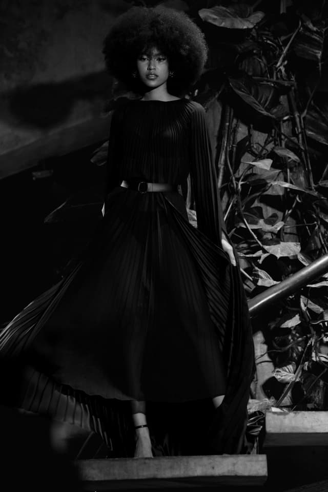 Black-and-white fashion portrait of a woman in a flowing dark gown standing on steps, framed by dramatic lighting and textured foliage