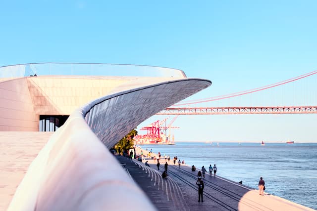 A modern architectural structure with a sweeping, curved design is situated by a waterfront People are walking along the promenade, and a large bridge spans the background under a clear blue sky