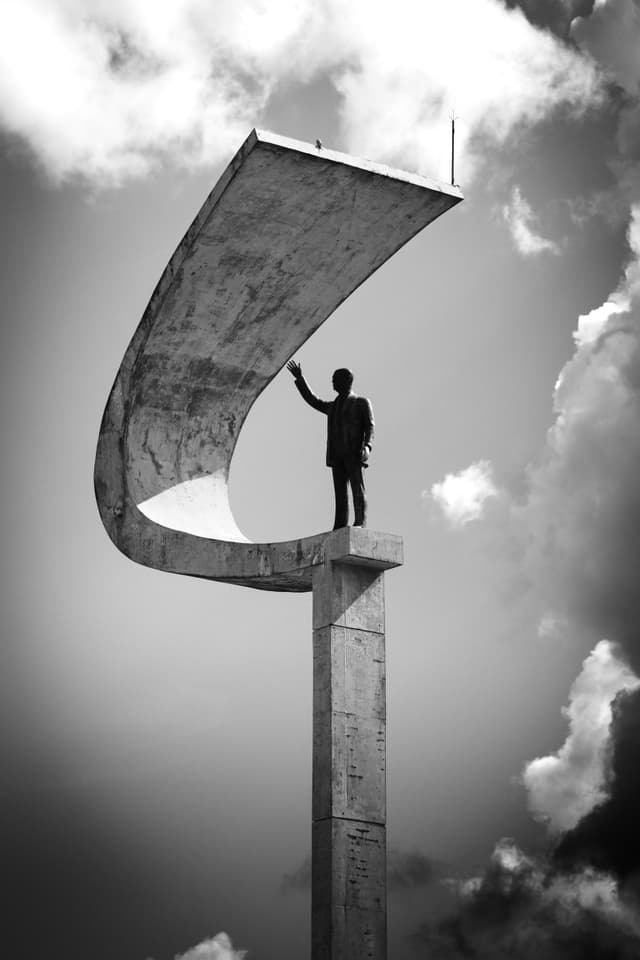 A black and white photograph of a tall, curved concrete structure with a silhouette of a person standing on top, set against a cloudy sky