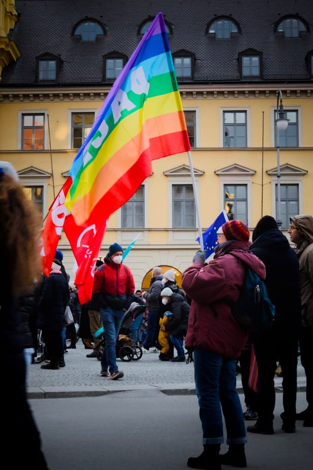 A group of people gathered outdoors, holding colorful flags, including a rainbow flag, in front of a building