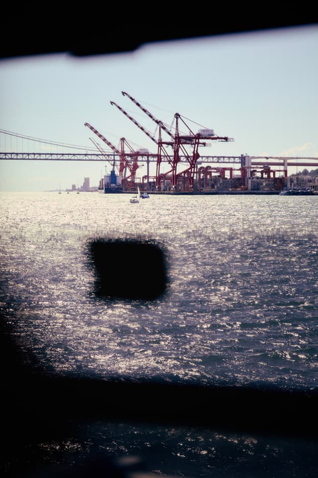 A waterfront scene with large red cranes, a bridge in the background, and sunlight reflecting on the water