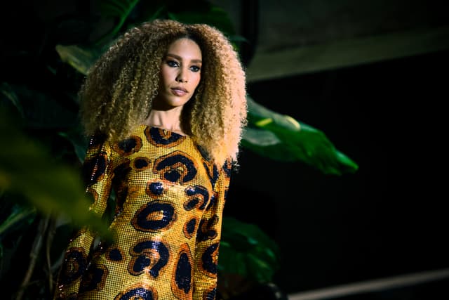 Studio portrait of a woman with curly hair wearing a leopard-print dress against a dark background with green leaves