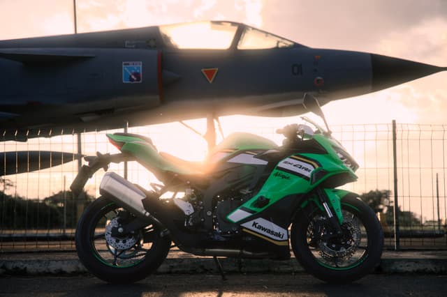 A green motorcycle parked beneath a fighter jet with the sun setting in the background