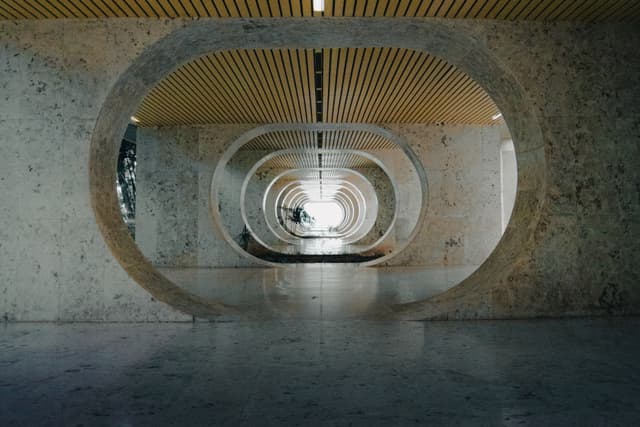 A modern architectural hallway with large circular openings and a reflective floor, leading to a bright light at the end