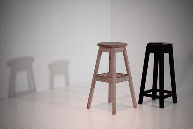 Two stools, one light brown and one black, are placed on a white surface with their shadows cast on the wall behind them