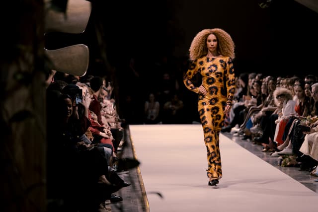 Fashion runway show with a model walking the catwalk in a leopard-print outfit while an audience watches from both sides