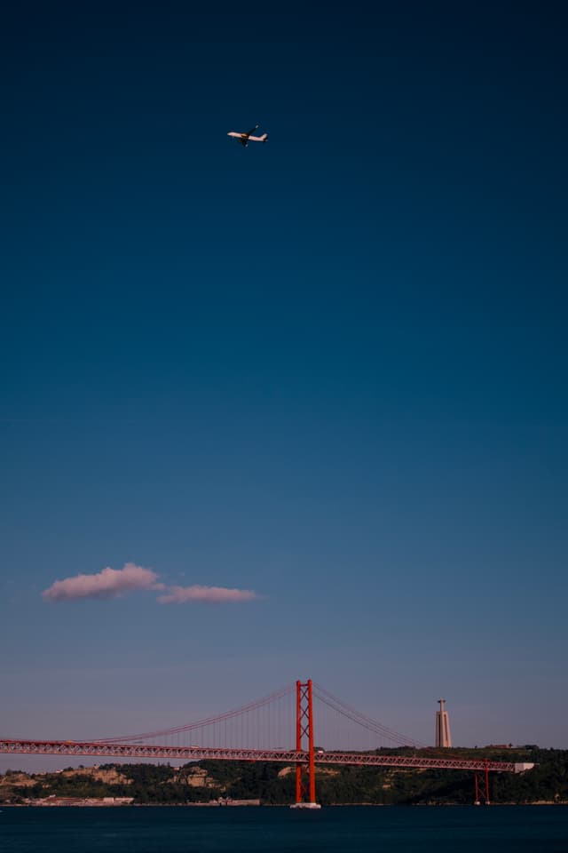 A red suspension bridge spans across a body of water with a plane flying above in a clear blue sky