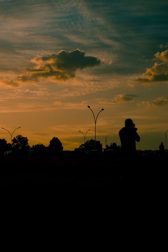 A silhouette of a person taking a photo against a vibrant sunset sky with scattered clouds and trees in the background