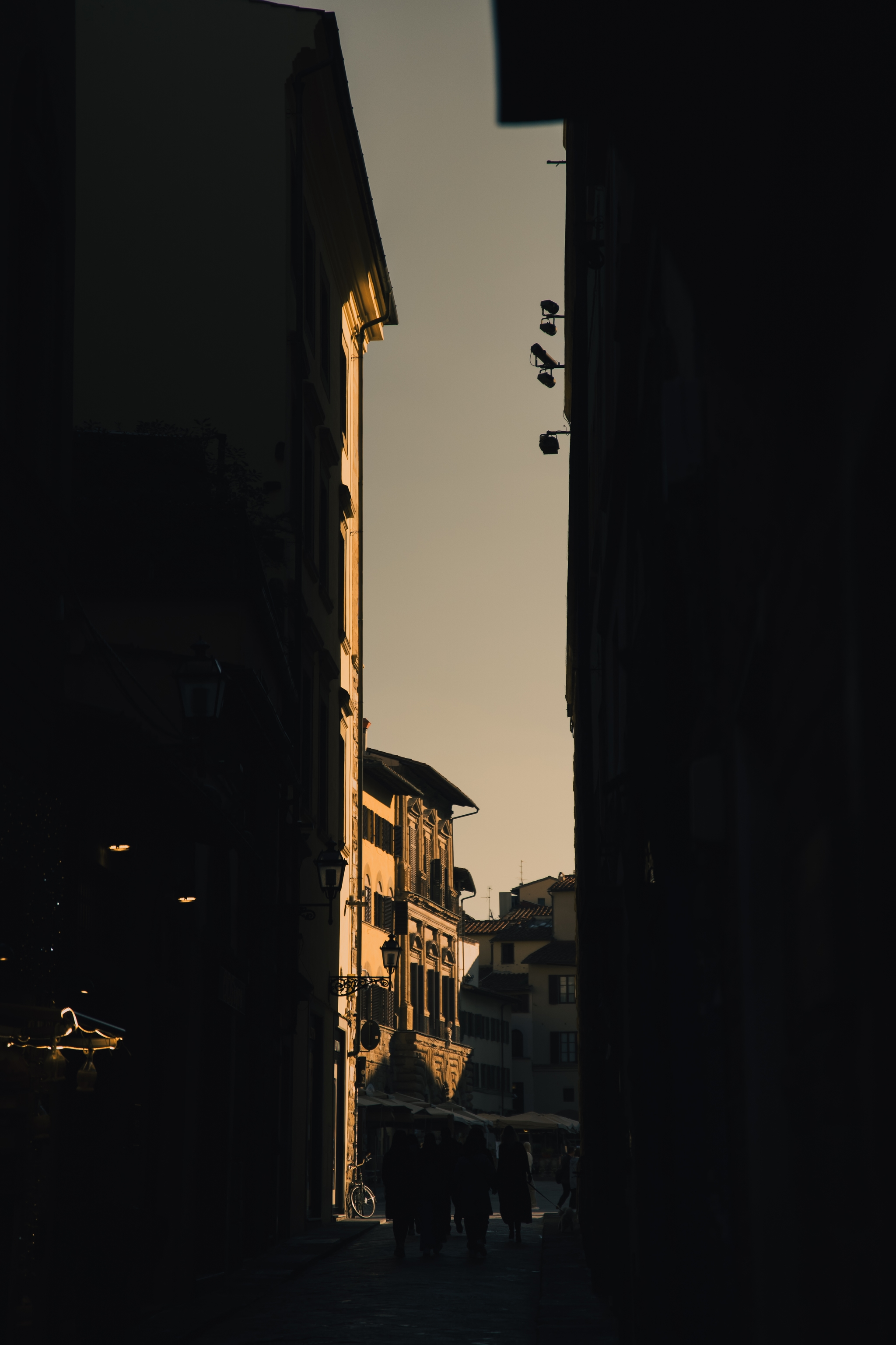 Narrow city alleyway with tall buildings and warm sunlight illuminating the far end with distant pedestrians