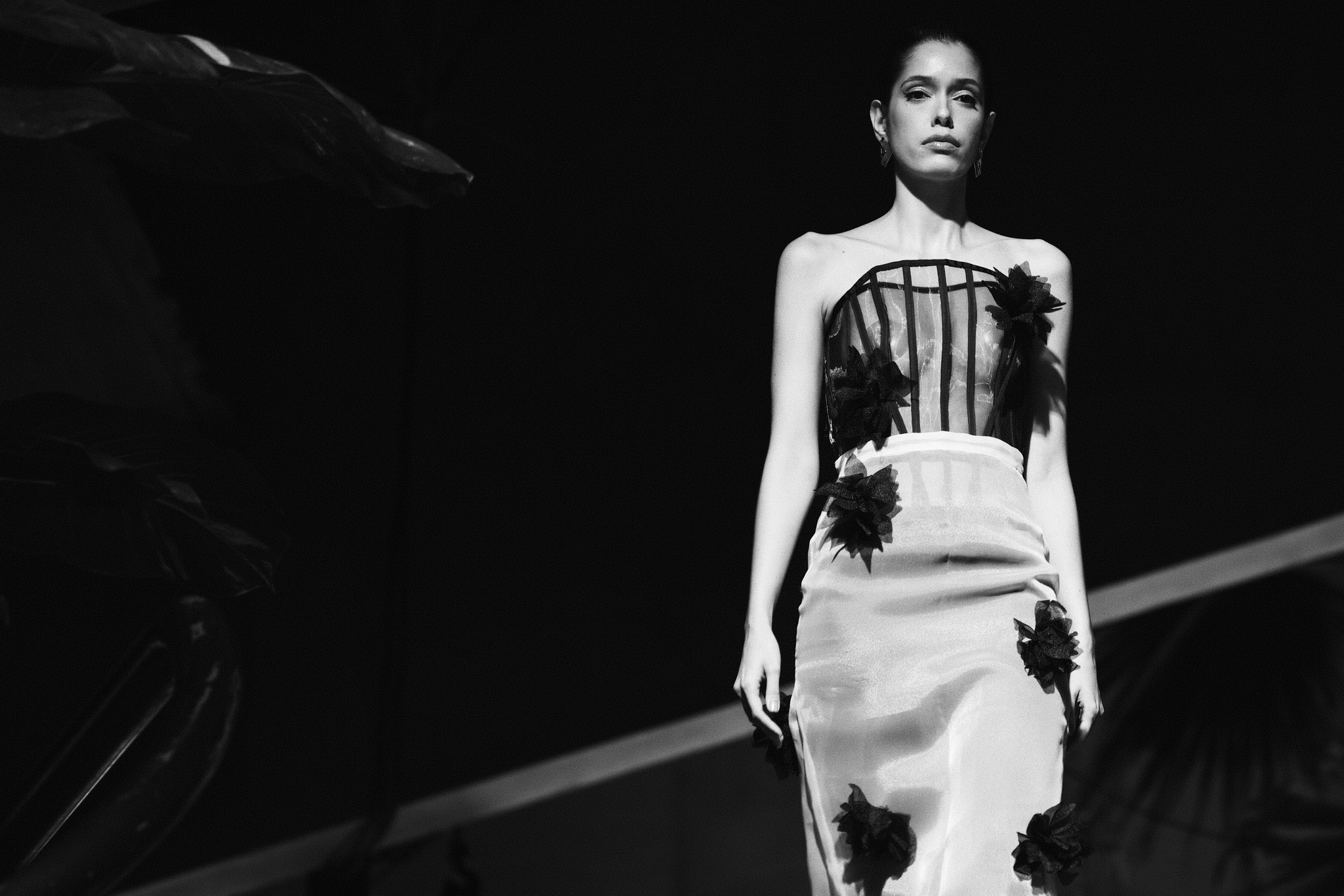 Black-and-white fashion runway scene with a model wearing a strapless corset-style dress walking under stage lighting