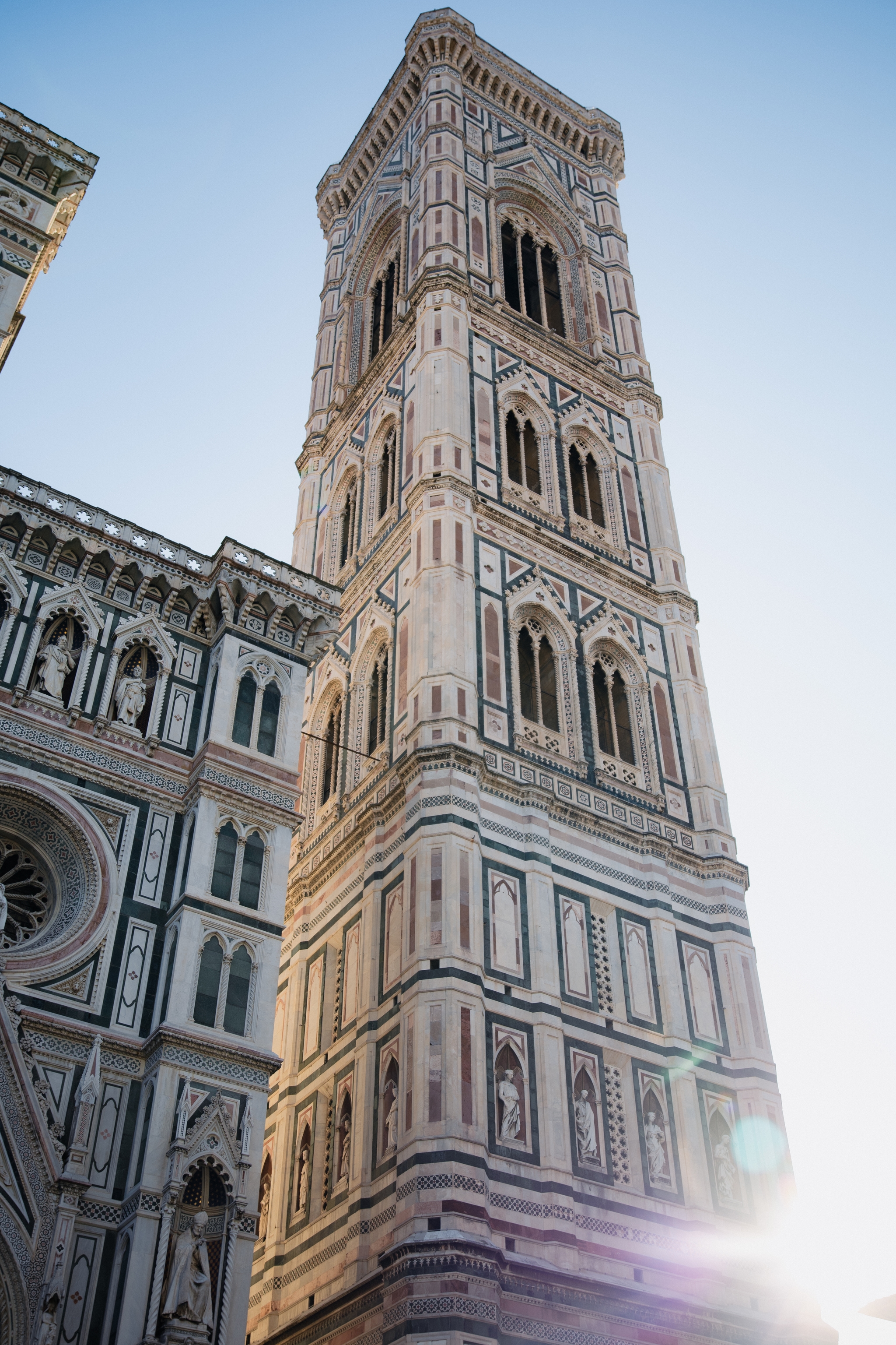 Low-angle view of Giotto’s Campanile beside the Florence Cathedral facade, with sun flare against a clear blue backdrop