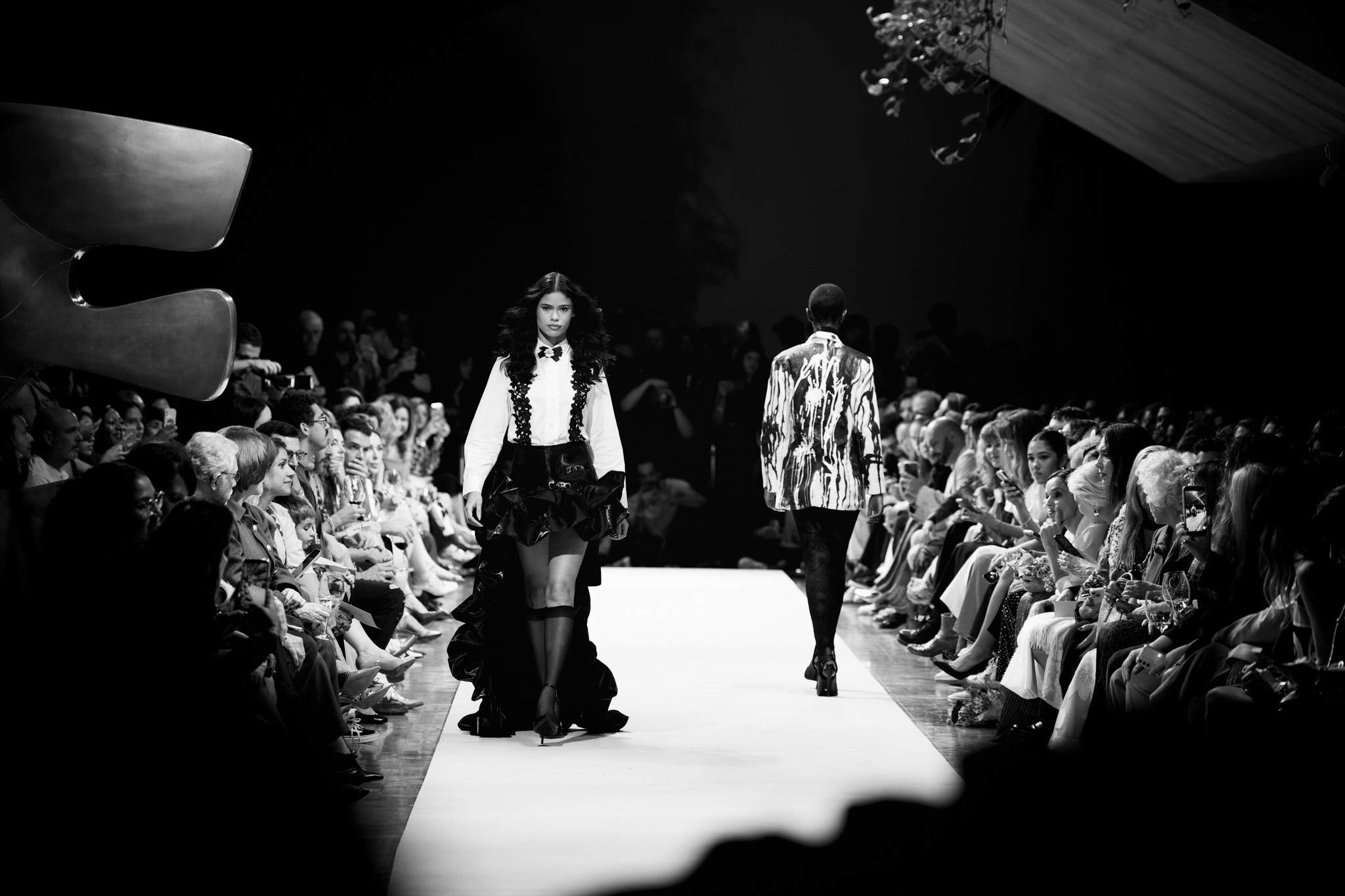 Black-and-white fashion runway scene with two models walking under spotlights as a seated crowd watches from both sides