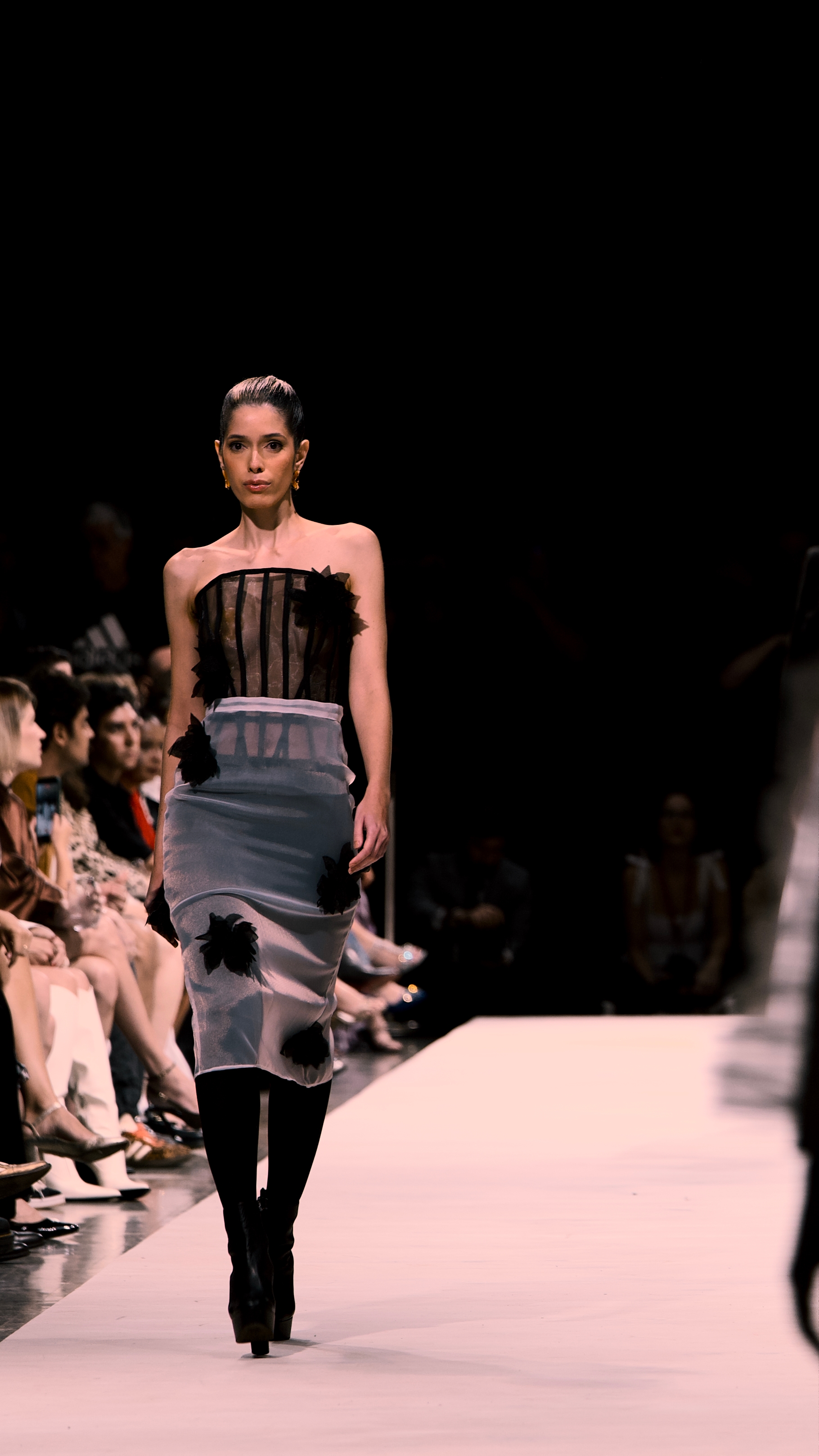 Fashion model walking down a runway in a strapless top and fitted skirt, with seated audience along the sides under dramatic stage lighting