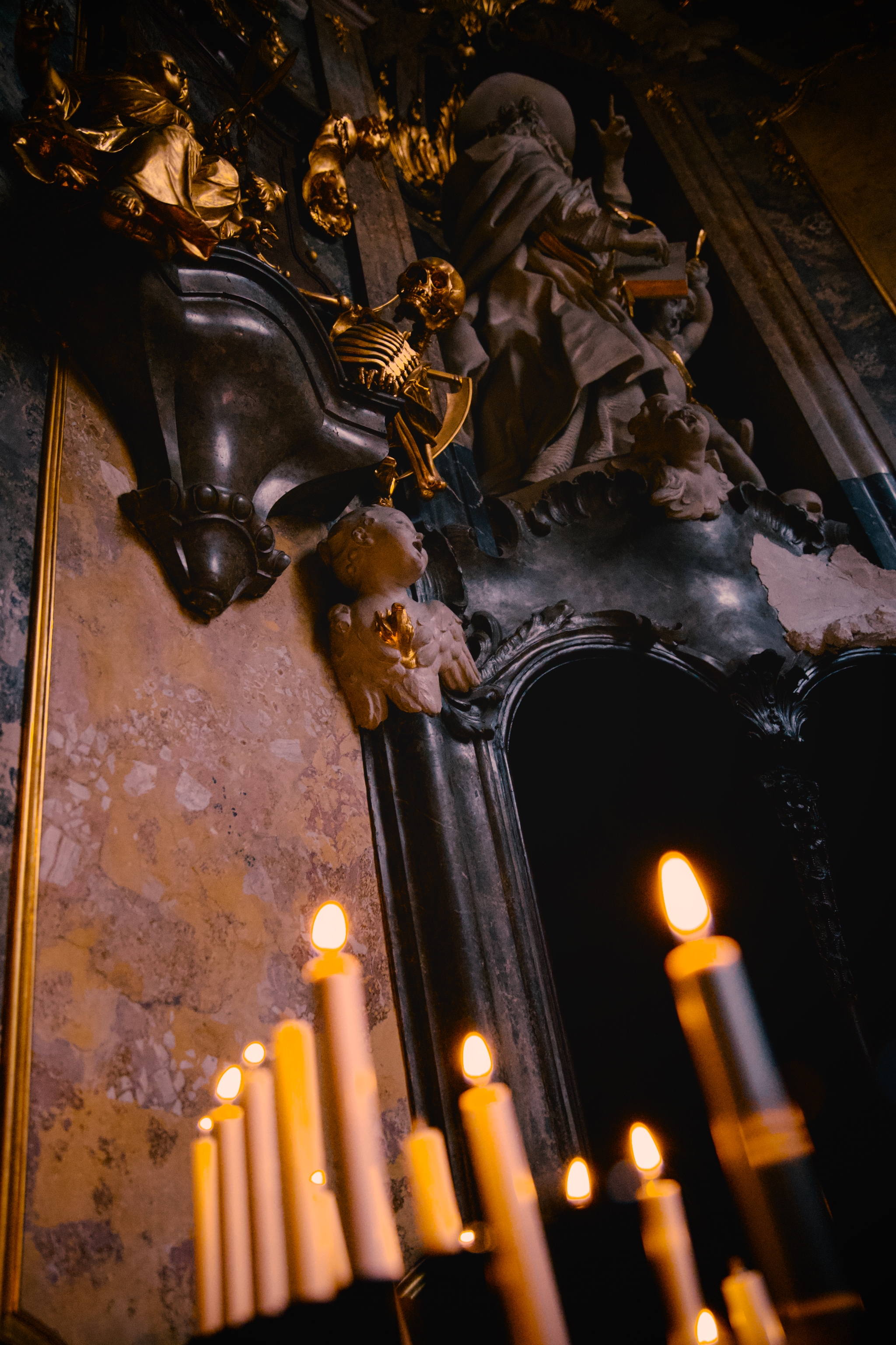 Lit candles in the foreground before an ornate, shadowed altar with carved figures and decorative stonework inside a dim interior