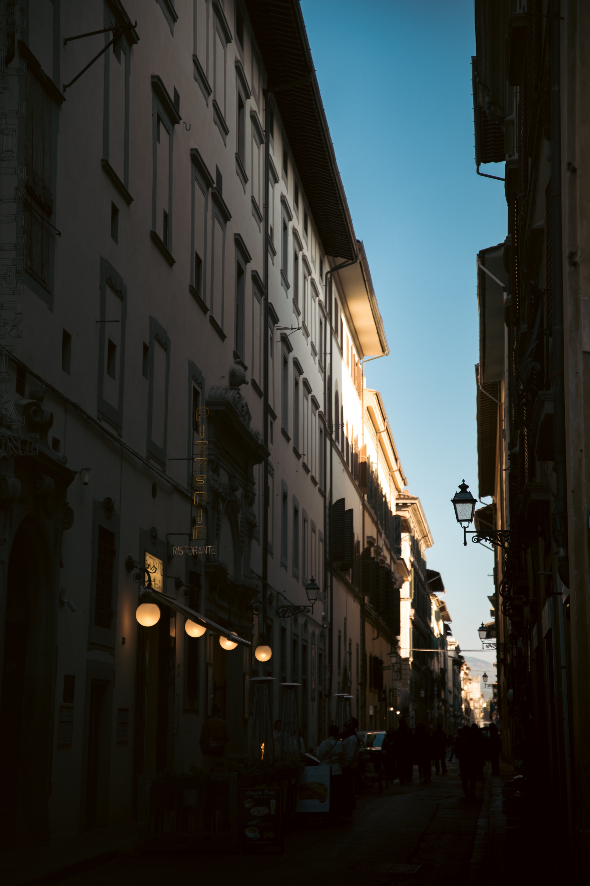 Narrow old-town street between tall buildings with streetlamps, backlit by low sun under a clear blue sky