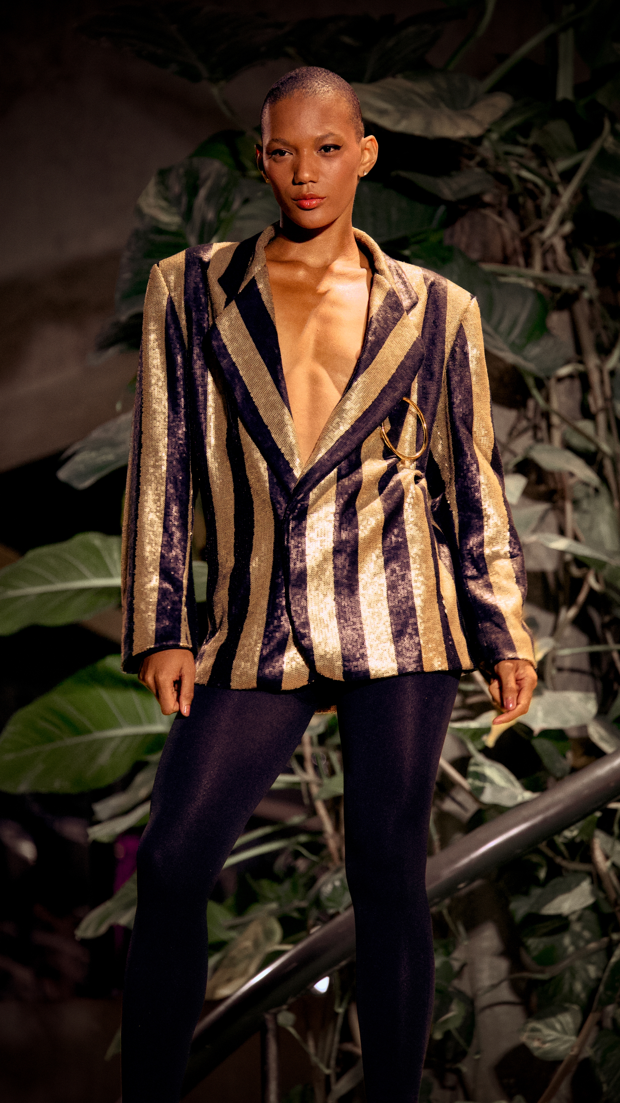 Fashion model on a runway wearing a striped blazer and dark leggings, standing against a leafy backdrop